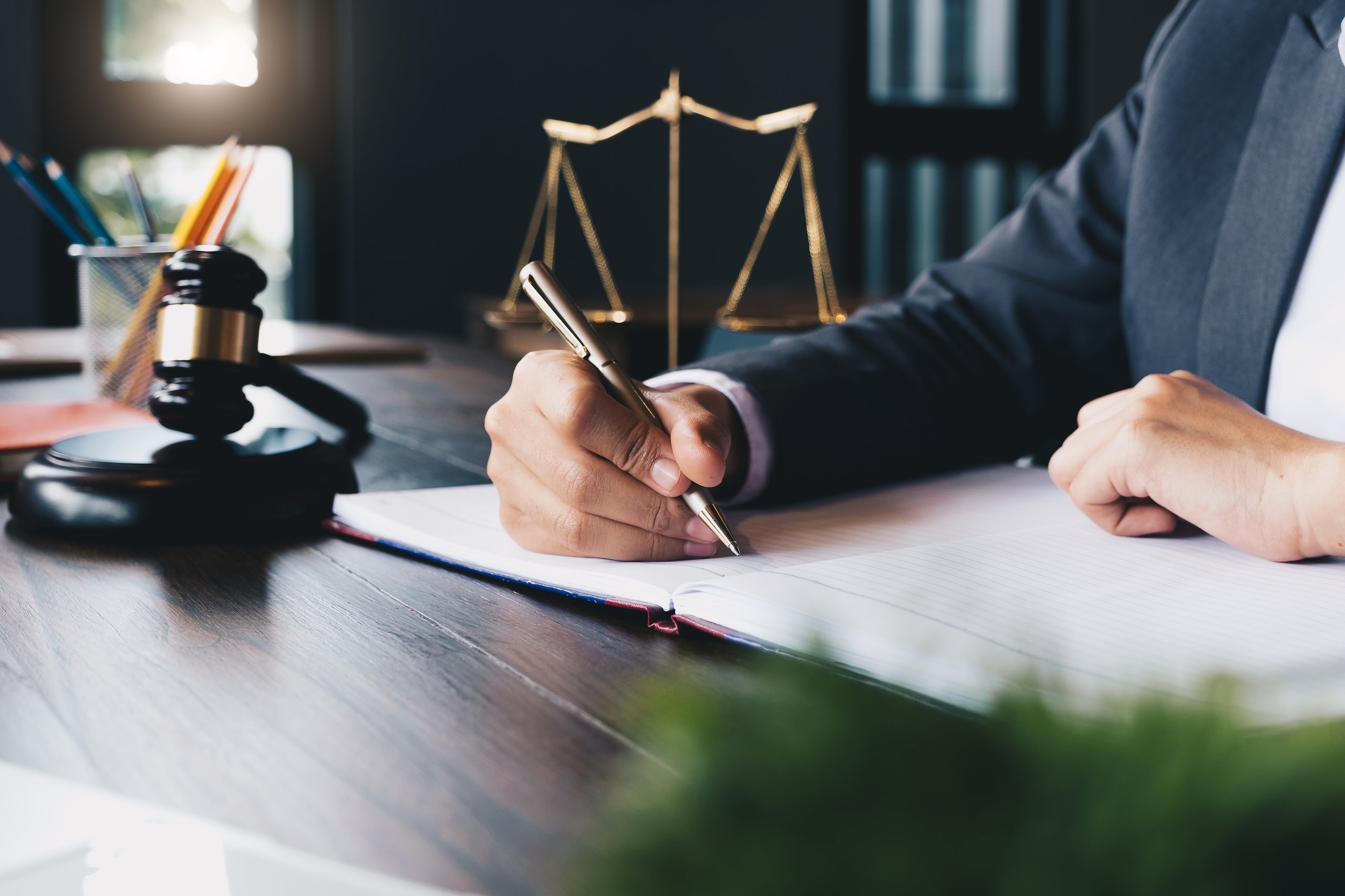 Person in business suit writing in an open notebook with a gavel and scales of justice nearby.