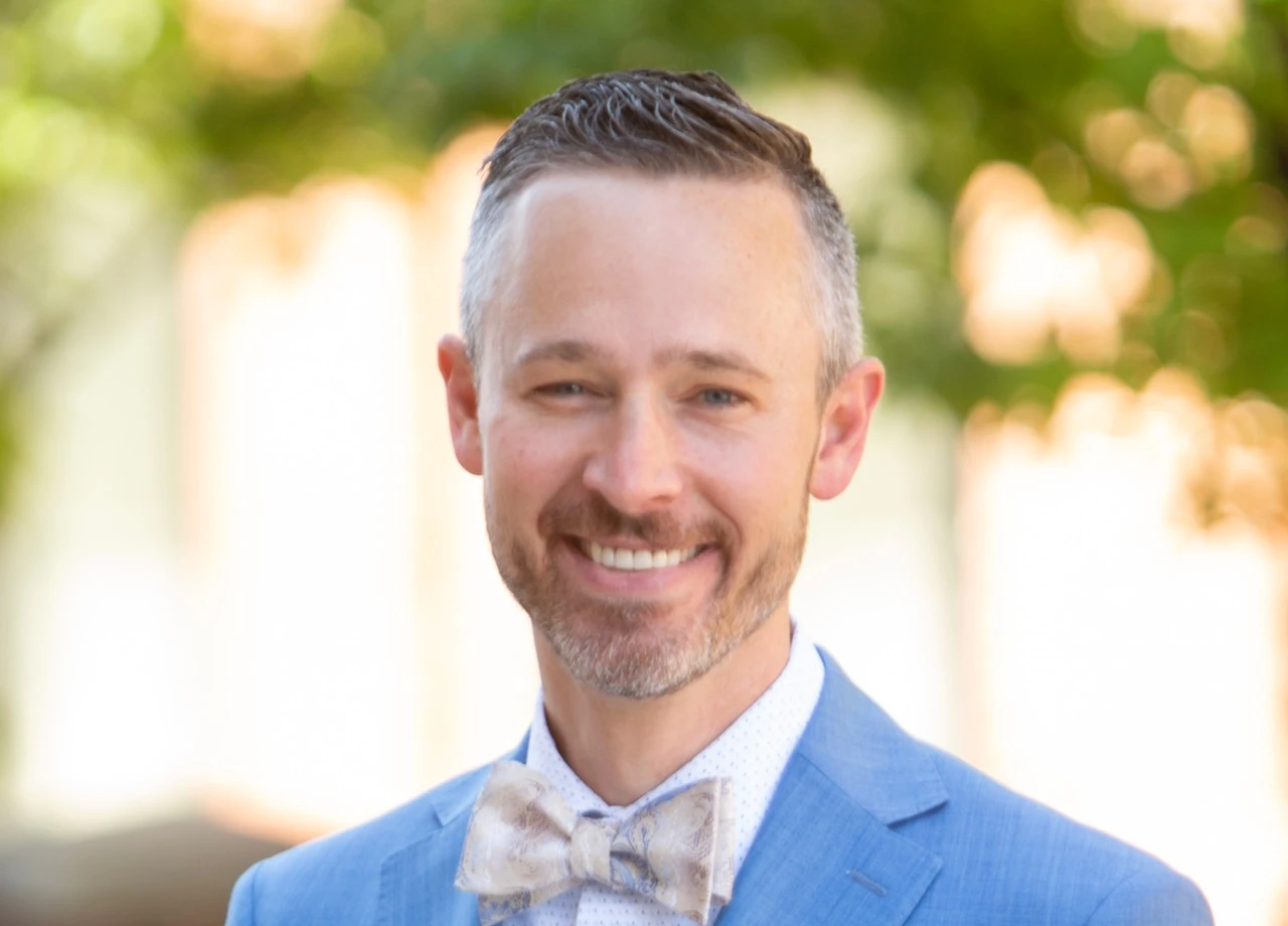 Smiling man with short gray hair and beard wearing a blue suit and patterned bow tie outdoors.