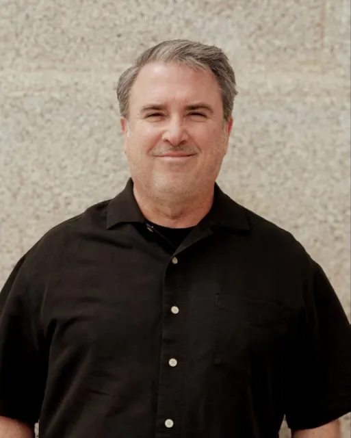 Middle-aged man with gray hair wearing a black collared shirt smiling against a light stone wall.
