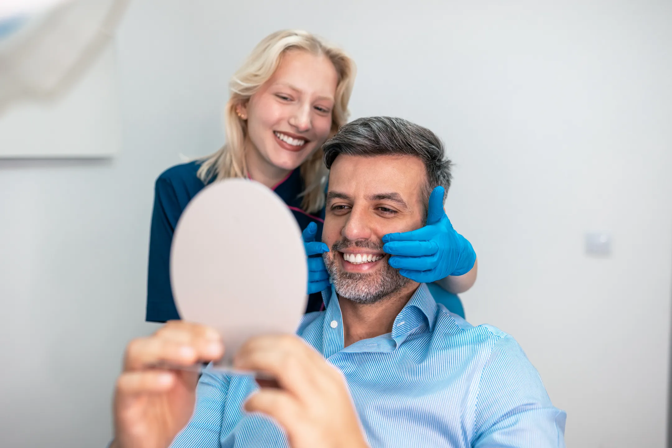 Dentist wearing blue gloves examines a smiling man's teeth as he holds a mirror.