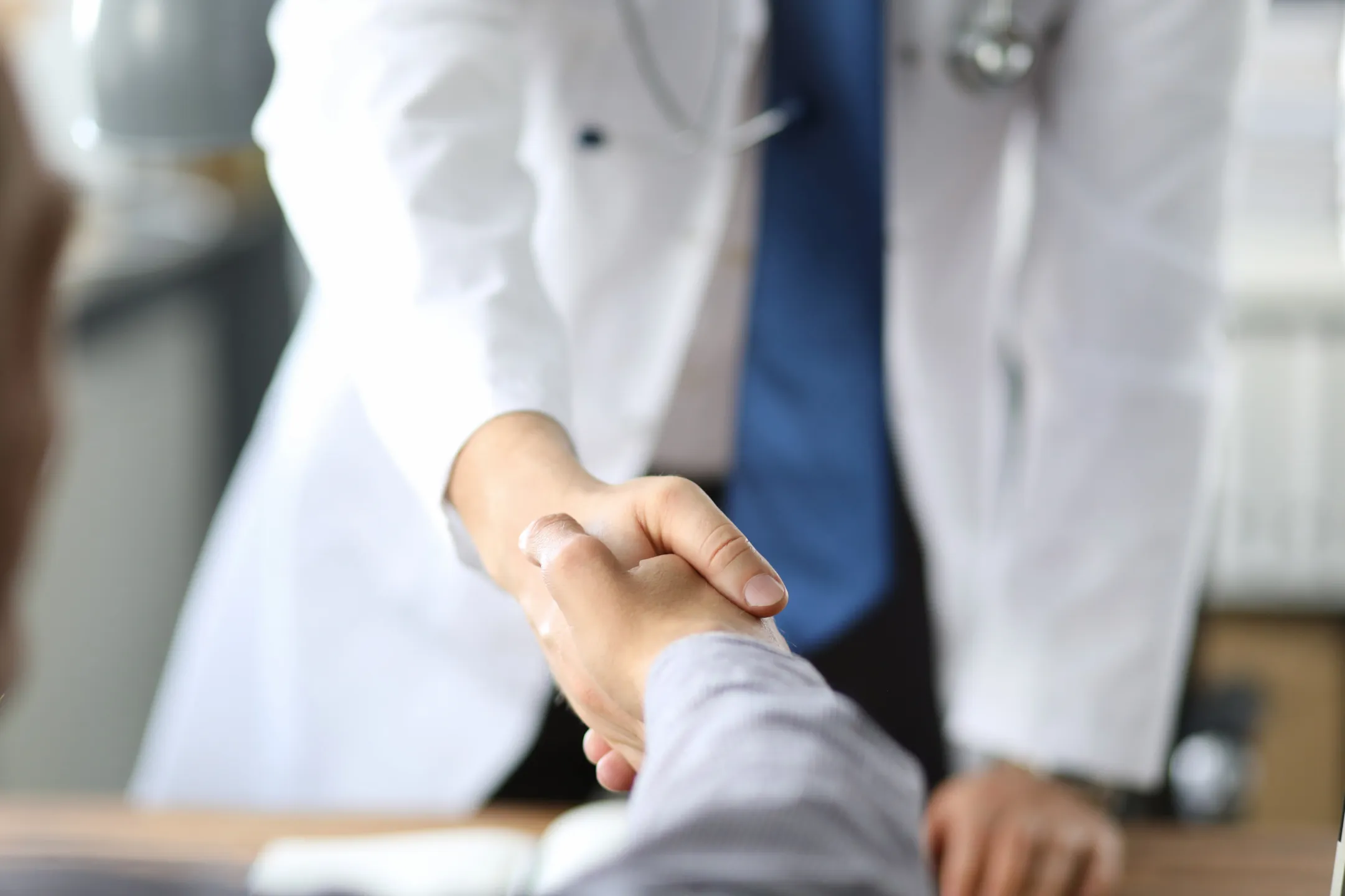 Doctor in white coat shaking hands with a patient across a desk in a medical office.
