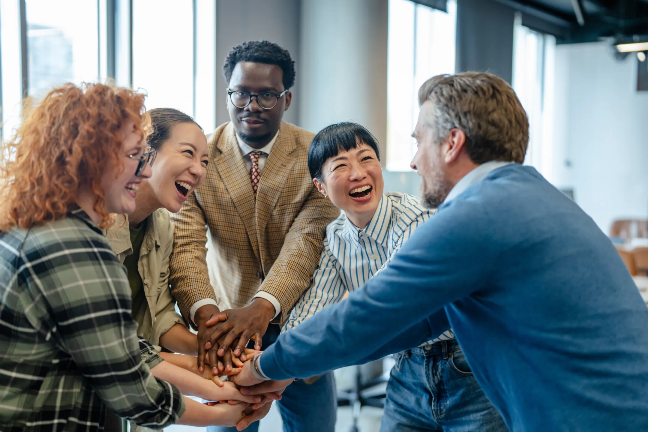 Diverse group of five people smiling and stacking their hands together in a teamwork gesture.