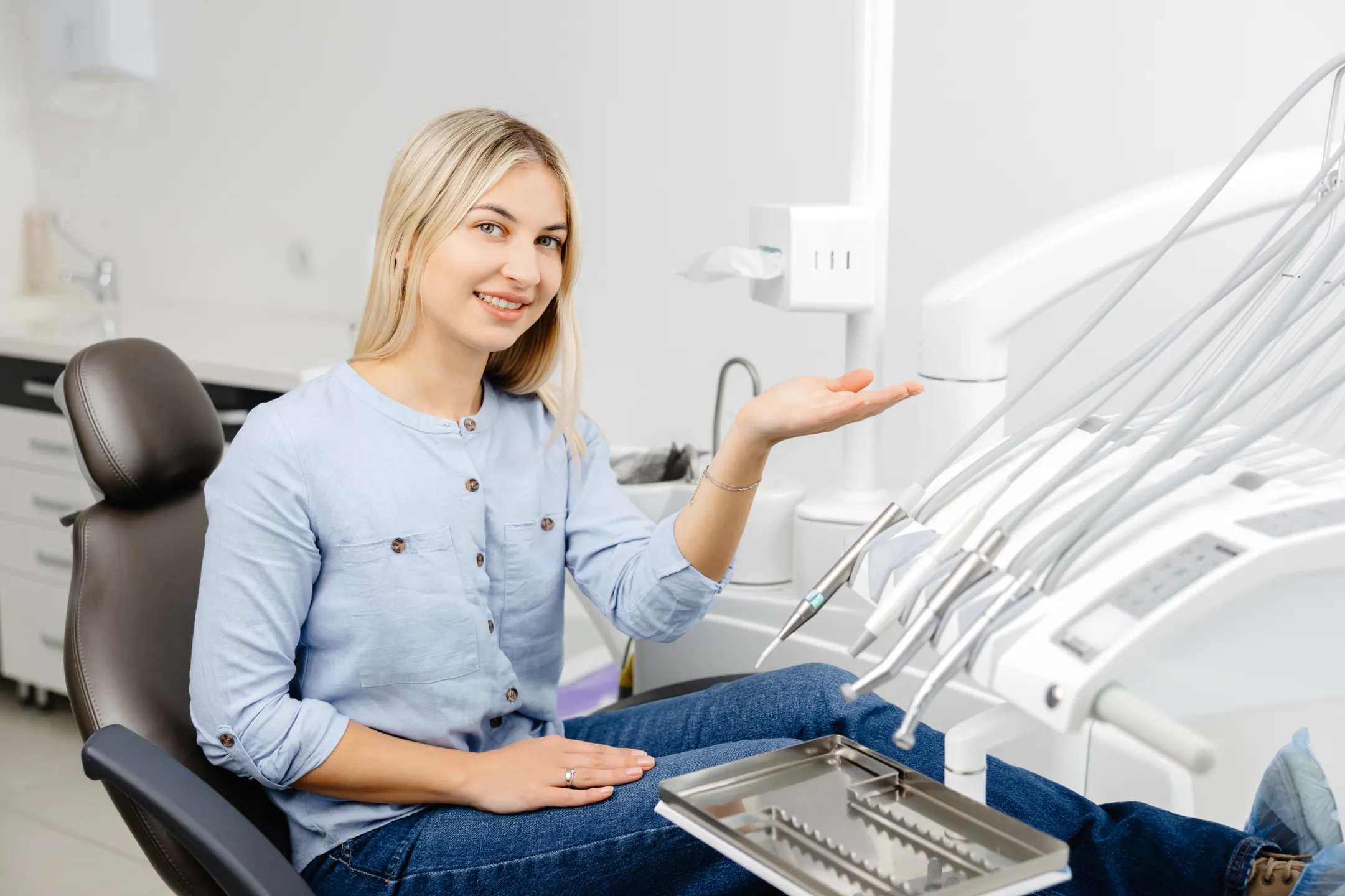 Smiling woman sitting in a dental chair in a clinic, gesturing toward dental equipment.