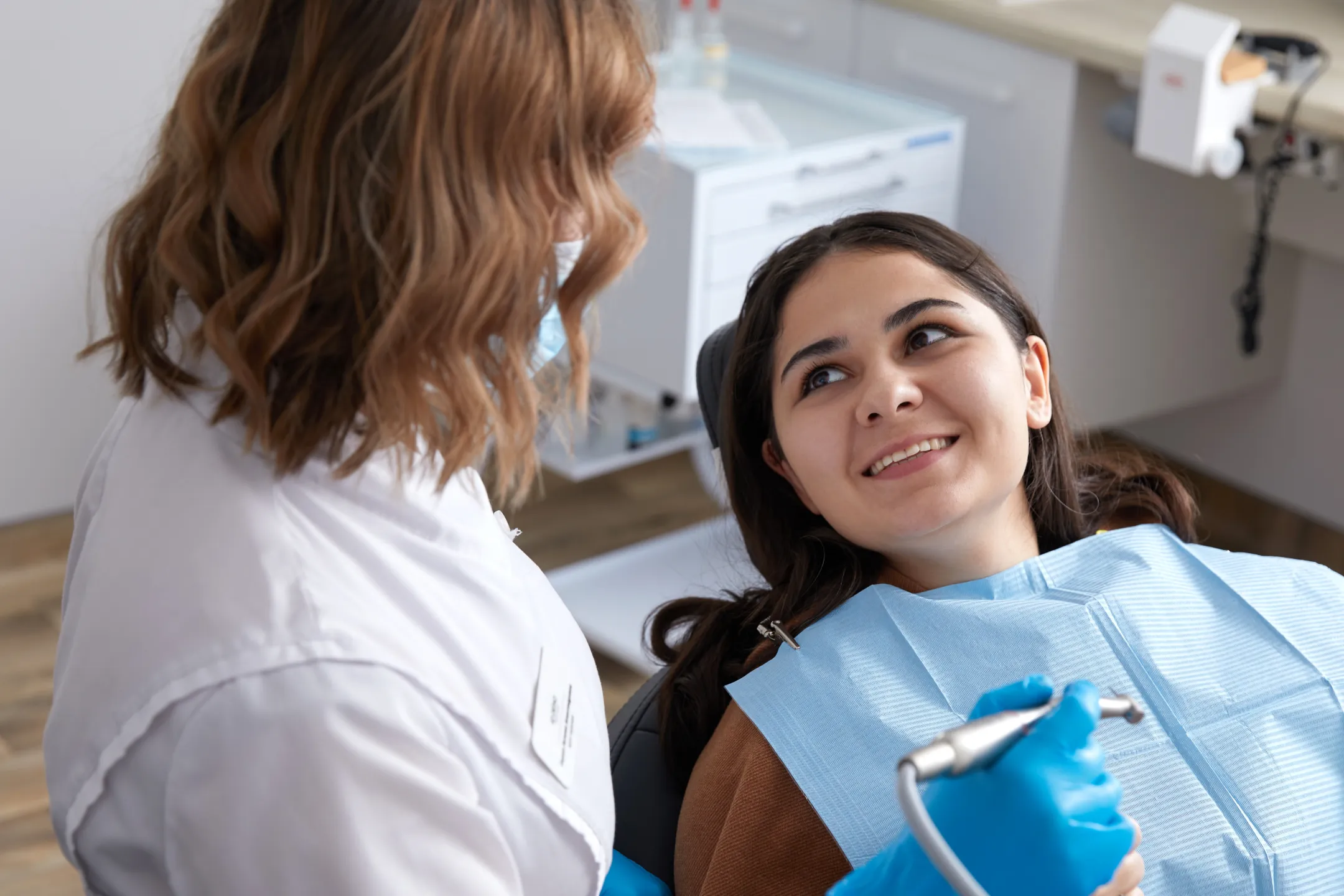 Smiling woman in dental chair looking at dentist holding a dental tool and wearing gloves.