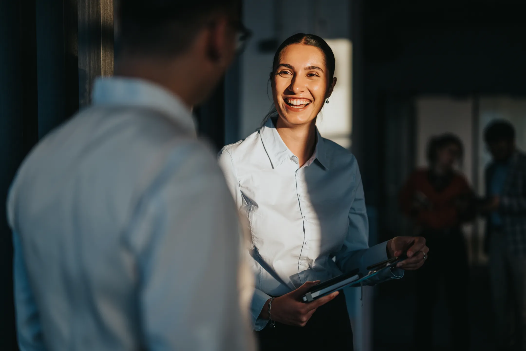 Smiling woman holding clipboard talks to a man in an office with people blurred in background.