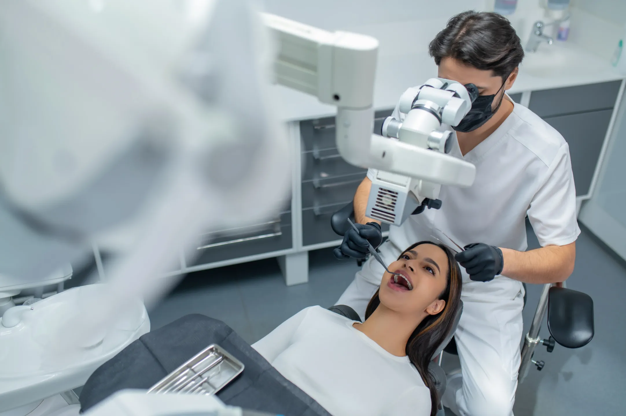 Dentist wearing gloves and mask examines female patient’s teeth using dental microscope in clinic.