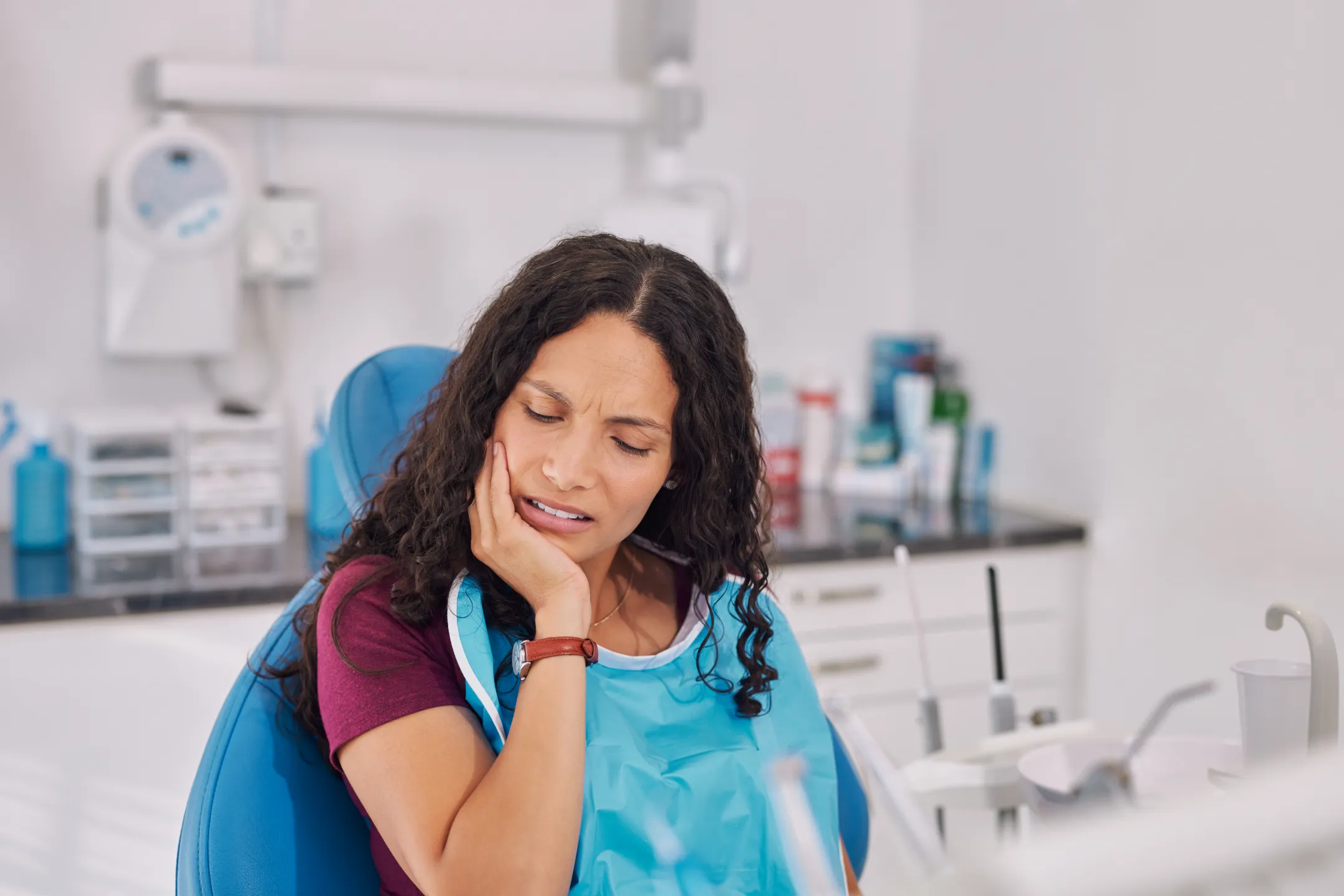 Woman in dental chair holding jaw in pain, wearing a dental bib in a clinic.