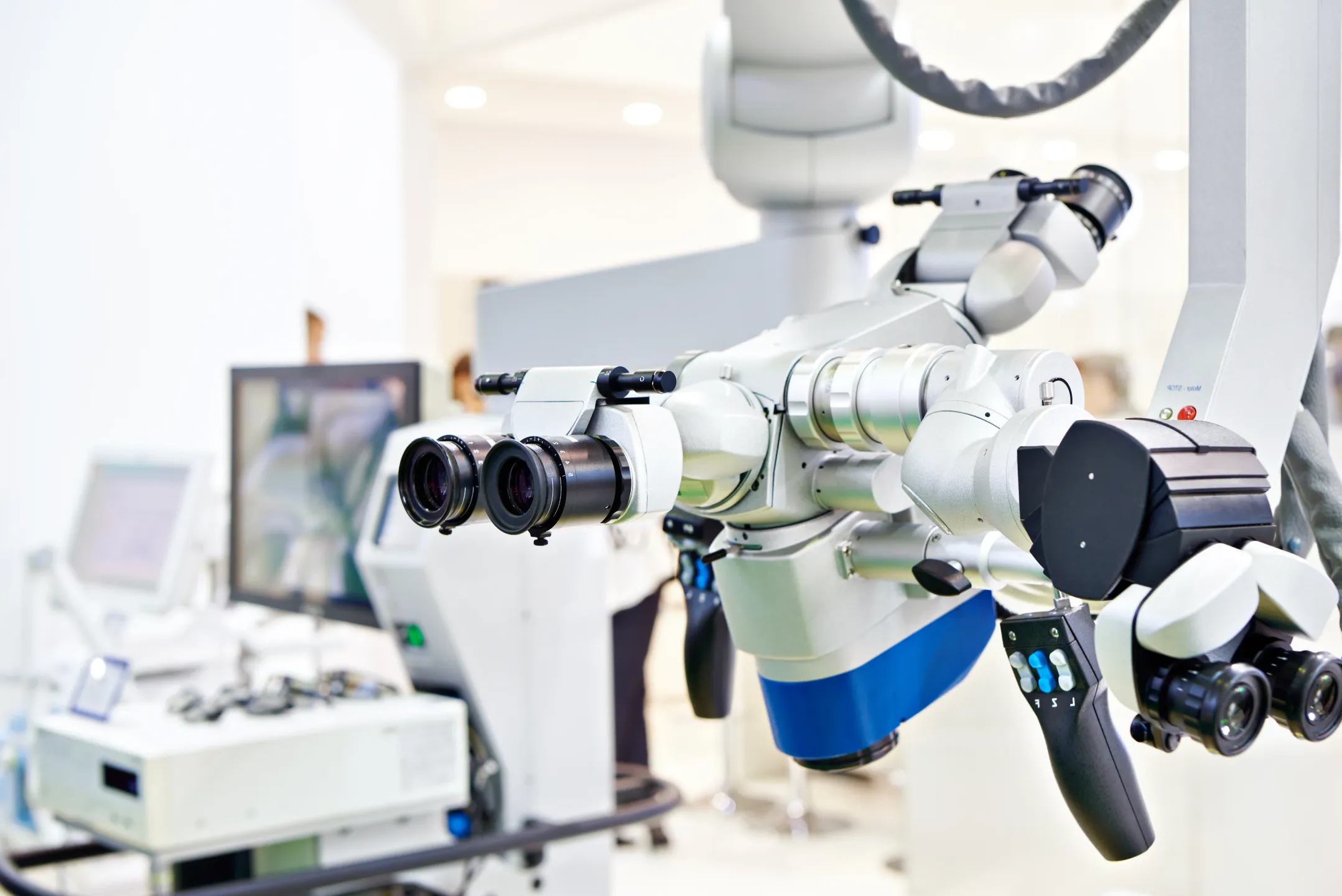 Close-up of a modern surgical microscope with multiple lenses and control handles in a medical setting.