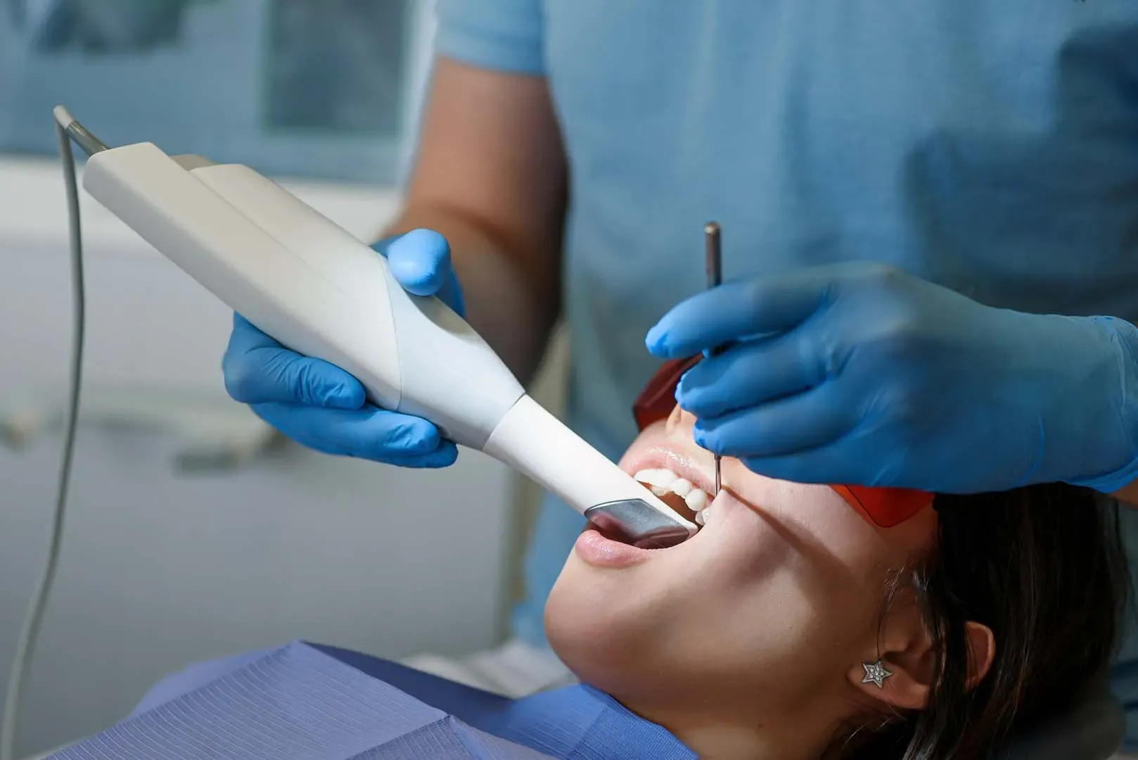 Dentist wearing blue gloves using dental scanner inside patient's open mouth with protective glasses.