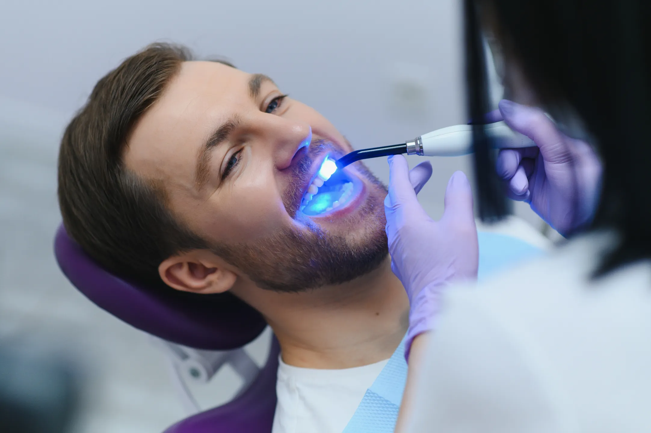 Dentist using a blue curing light to harden dental resin inside a male patient's mouth.