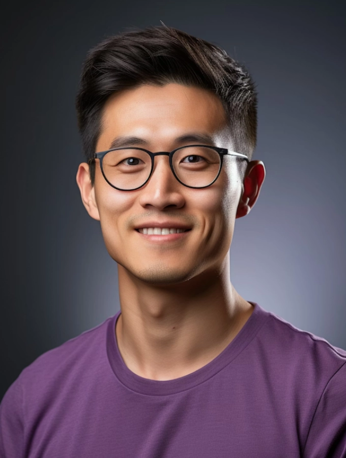 Smiling young man with short dark hair wearing round black glasses and a purple shirt against a dark gray background.