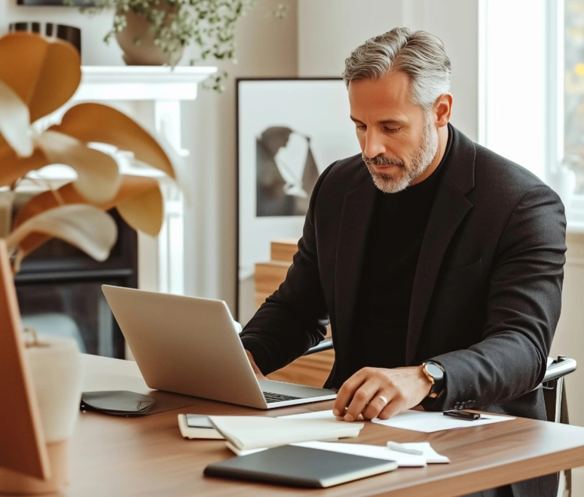 Middle-aged man with gray hair and beard working at a desk with a laptop and papers in a bright, modern room.