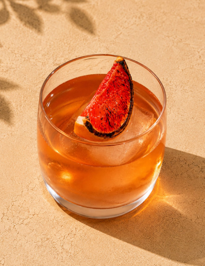 Glass of amber-colored cocktail with a large ice cube and a charred grapefruit wedge garnish.