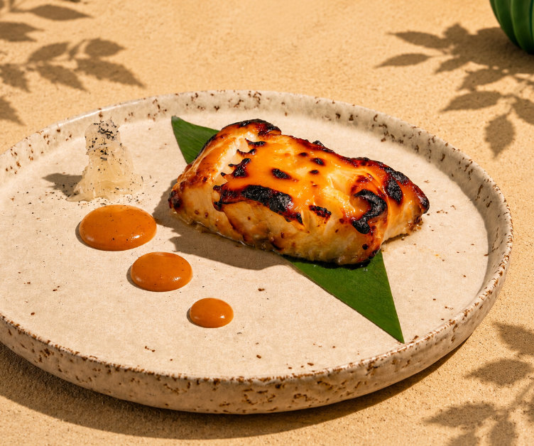Grilled fish fillet on a green leaf with three sauce drops and a translucent garnish on a rustic plate.