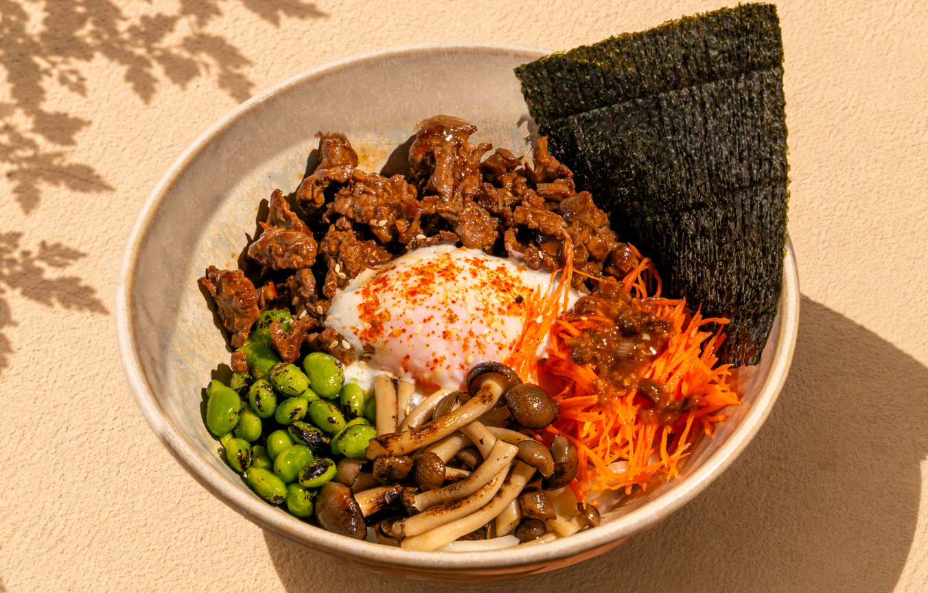 Bowl with seasoned beef, grilled mushrooms, edamame, shredded carrots, soft egg, and seaweed sheets.