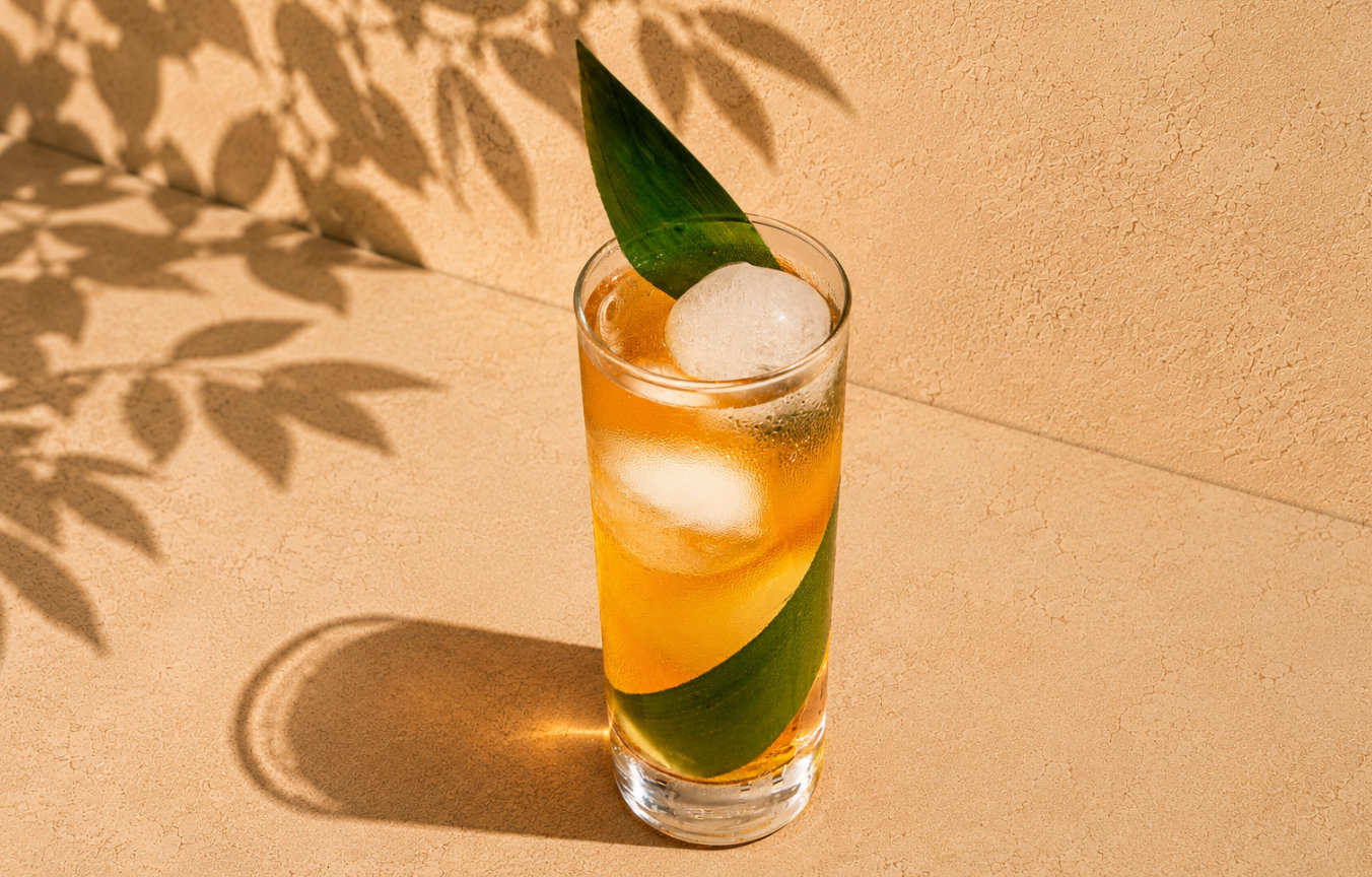 Tall glass with amber drink, ice balls, and a large green leaf garnish on beige surface.