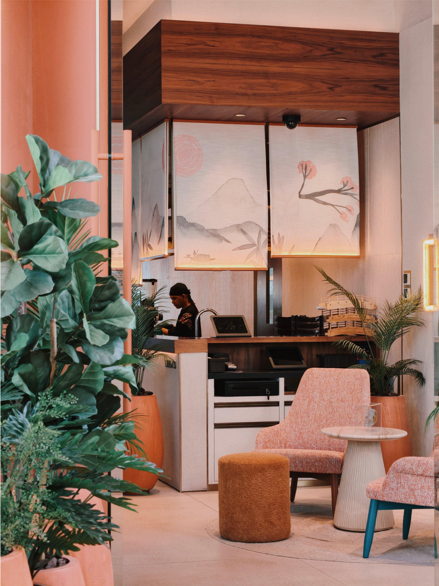 Modern reception with pink chairs, wooden counter, large plants, and a person working behind the desk.