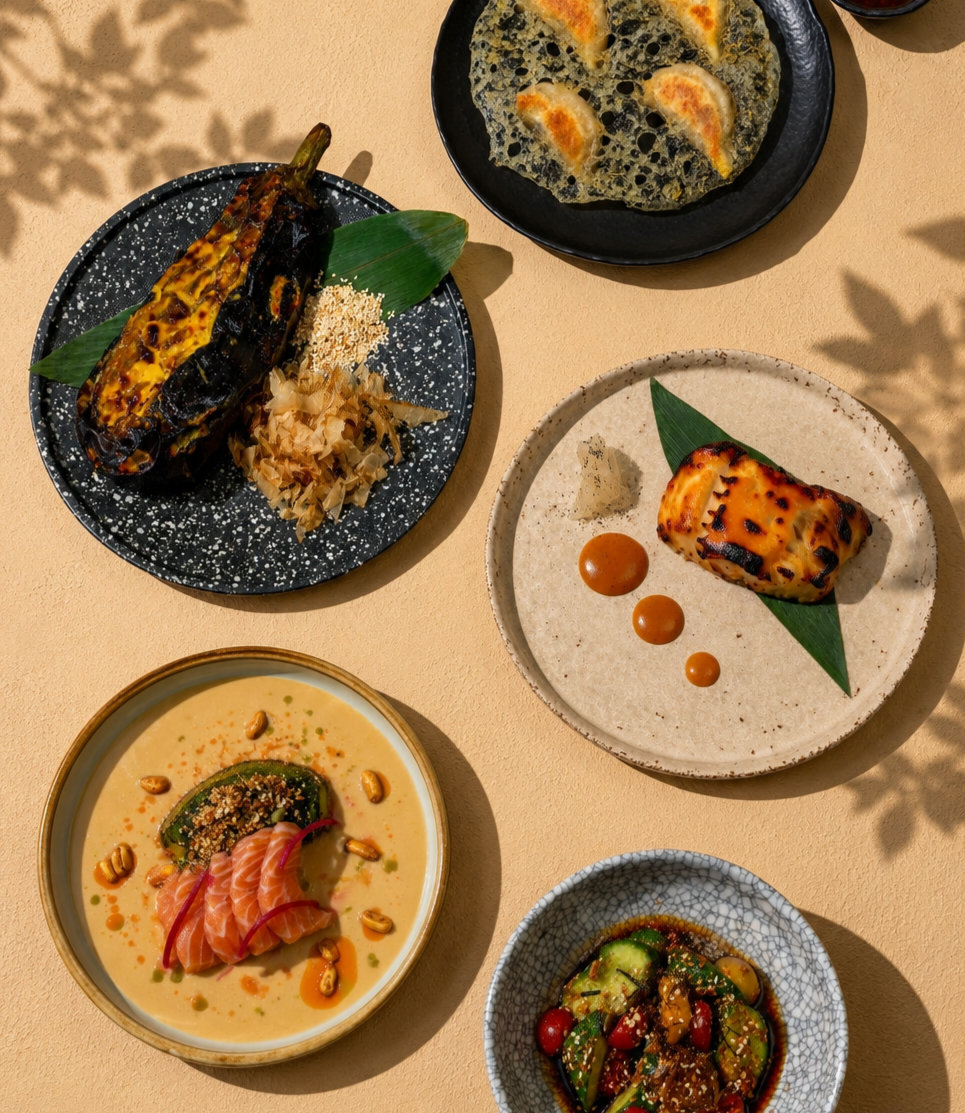 Overhead view of five plated gourmet dishes including grilled eggplant, dumplings, fish, salmon, and cucumber salad.