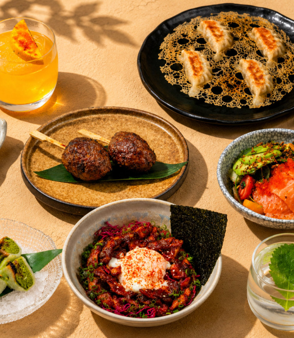 Table with Japanese dishes including grilled dumplings, skewers, poke bowl, green rolls, and two drinks.