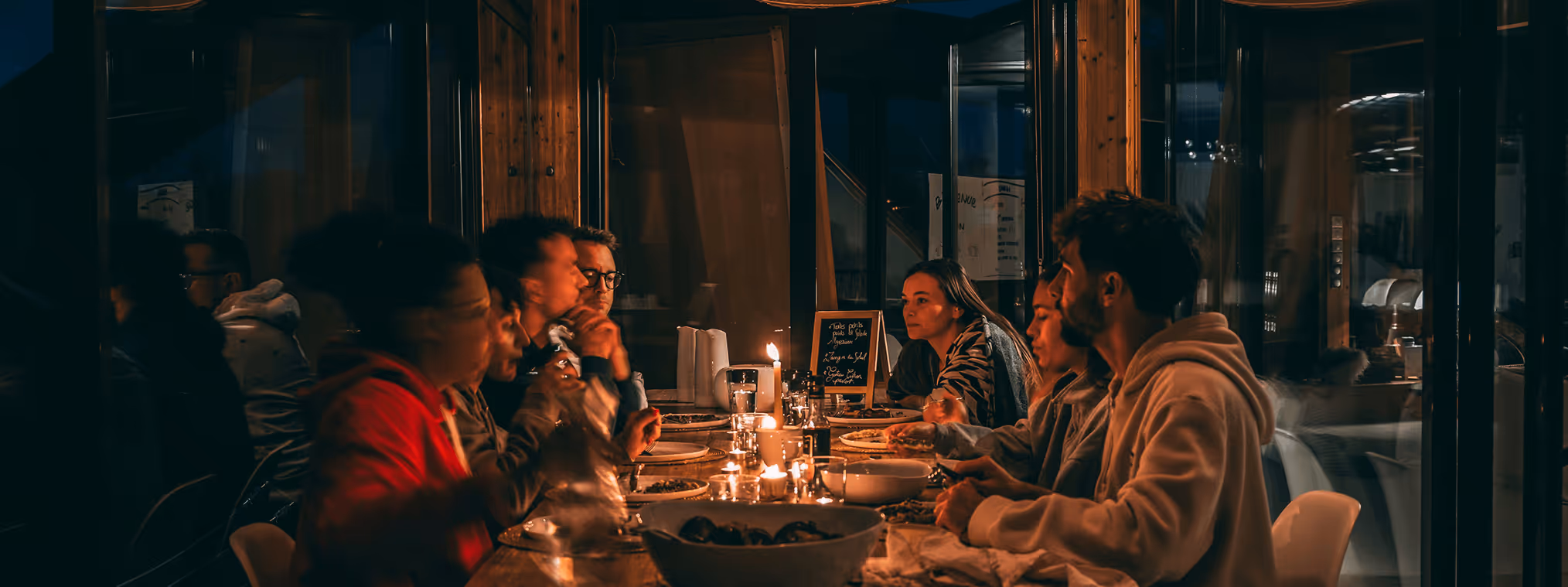 Group of friends having a candlelit dinner indoors around a wooden table with food and drinks.