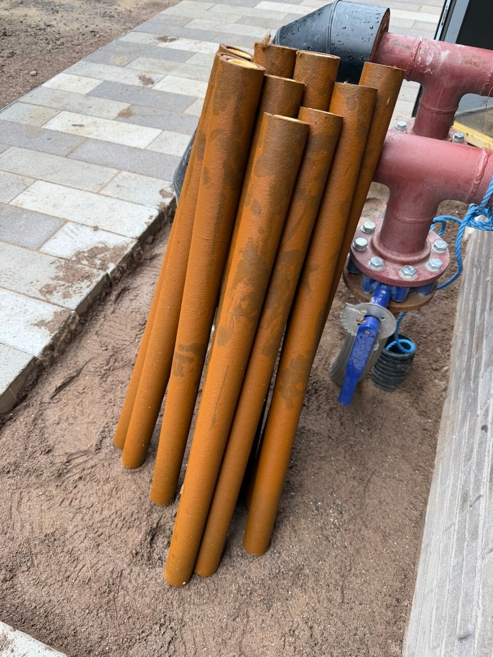 Cluster of rust-colored metal pipes leaning against a red valve on sandy ground beside tiled pavement.