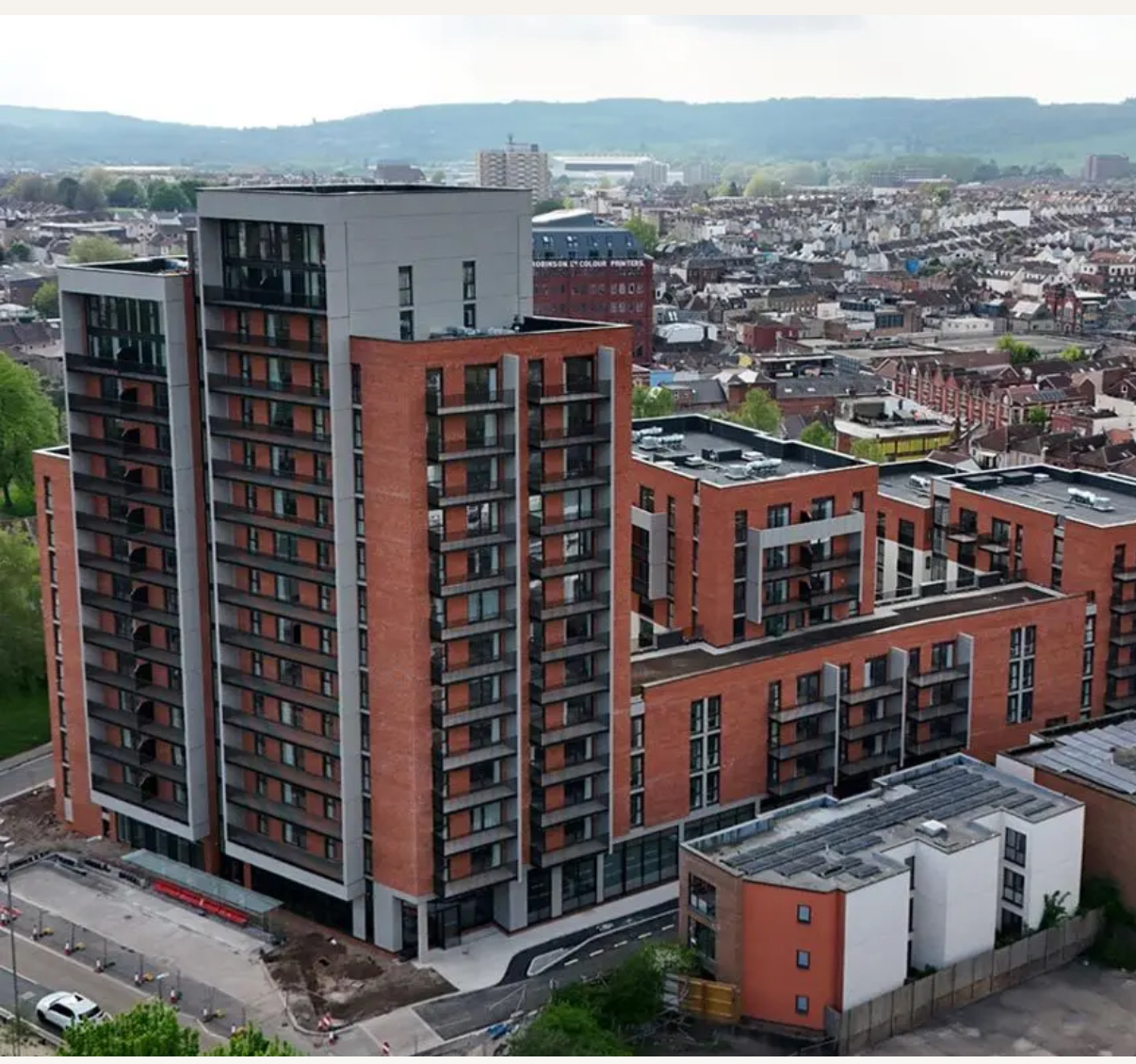 Modern multi-story apartment complex with balconies surrounded by a cityscape and distant hills.