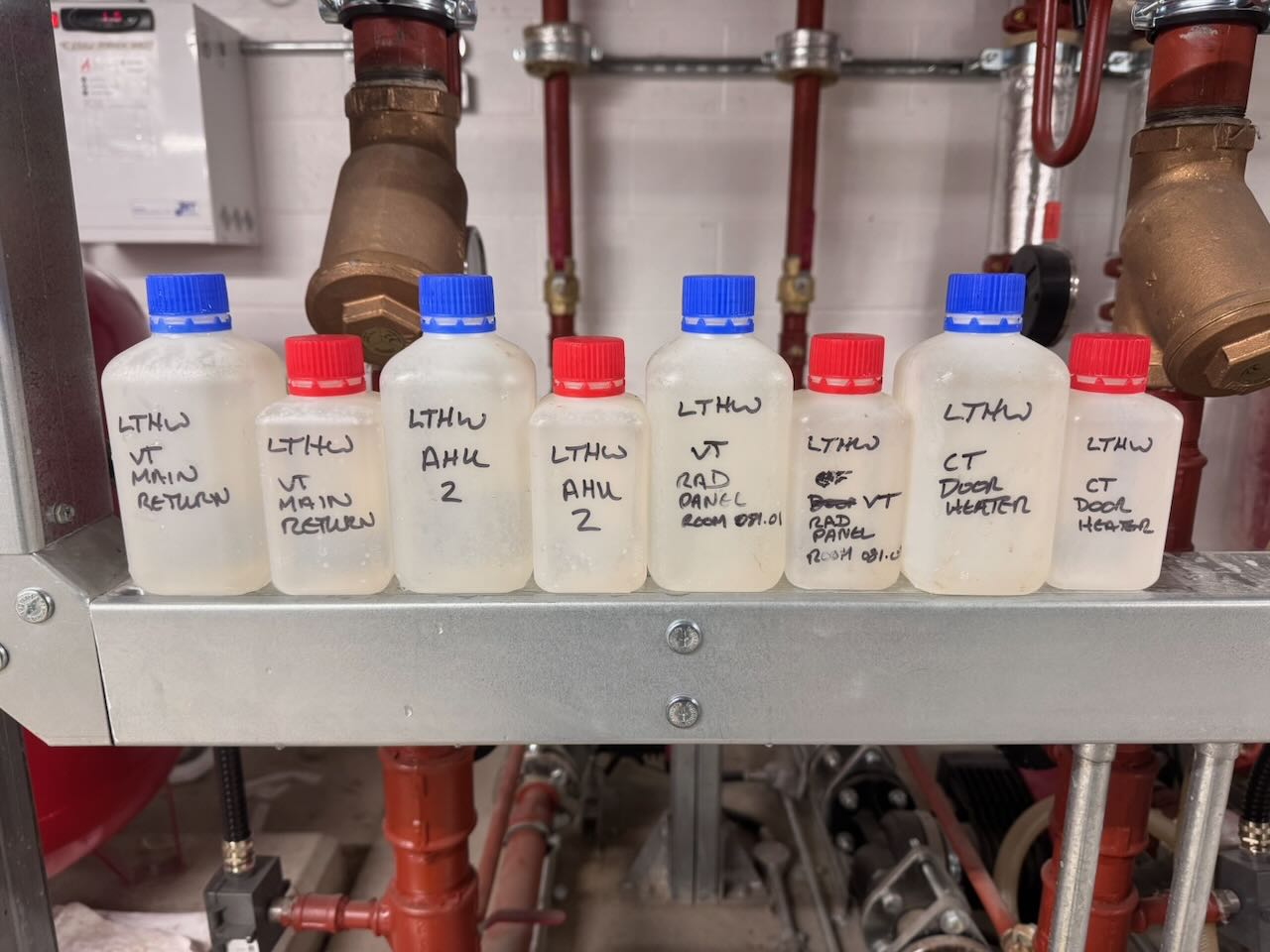 Eight small plastic bottles with red and blue caps labeled with handwritten text on a metal shelf in a mechanical room.