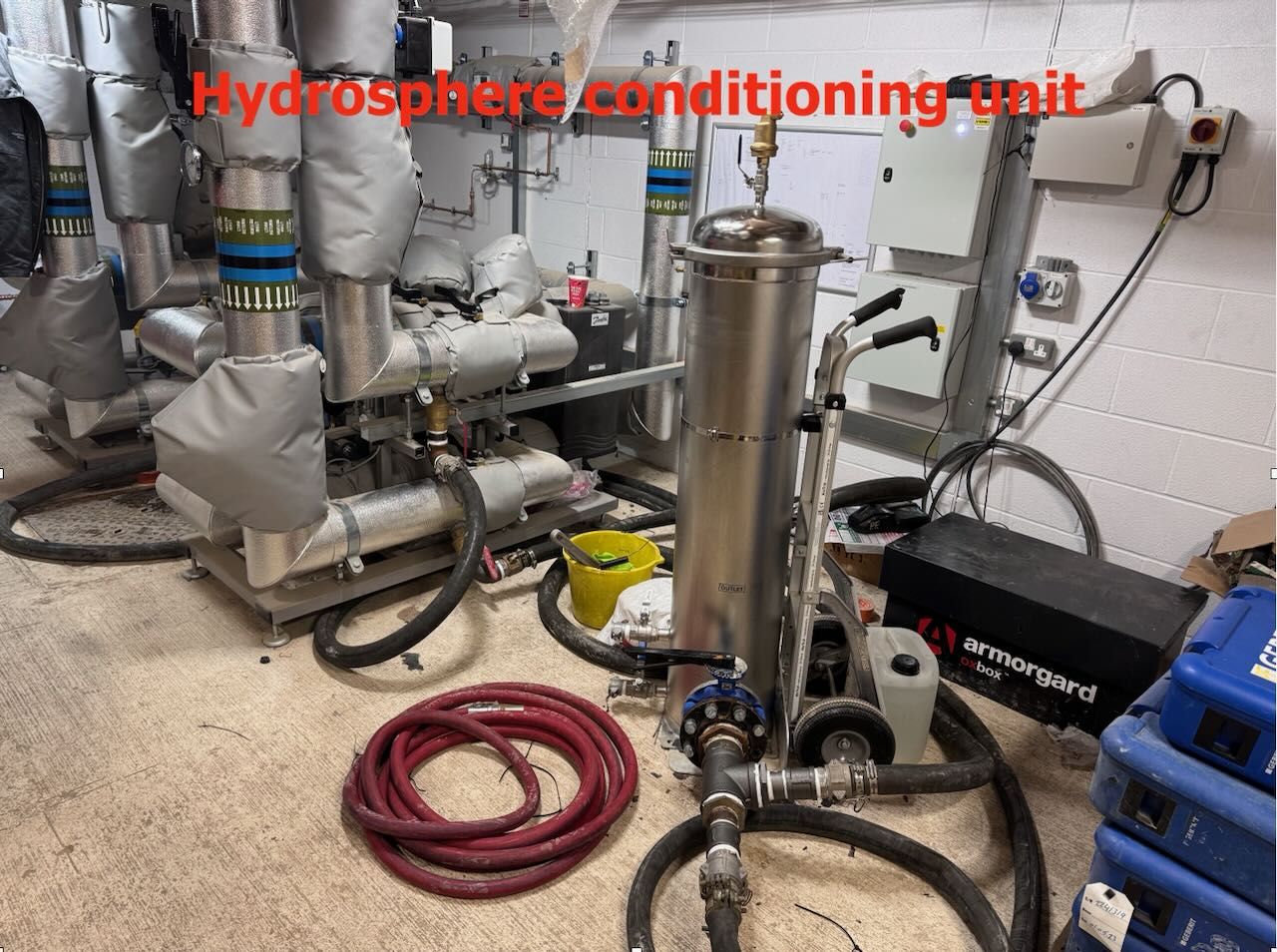Industrial room with insulated pipes, a silver hydrosphere conditioning unit, and coiled red hose on the floor.