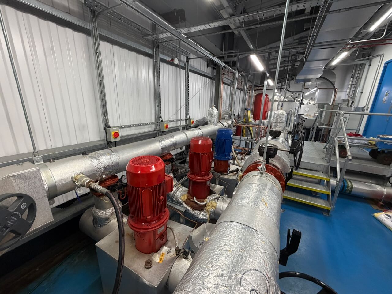 Industrial mechanical room with insulated pipes, red and blue pump motors, and metal platform stairs.
