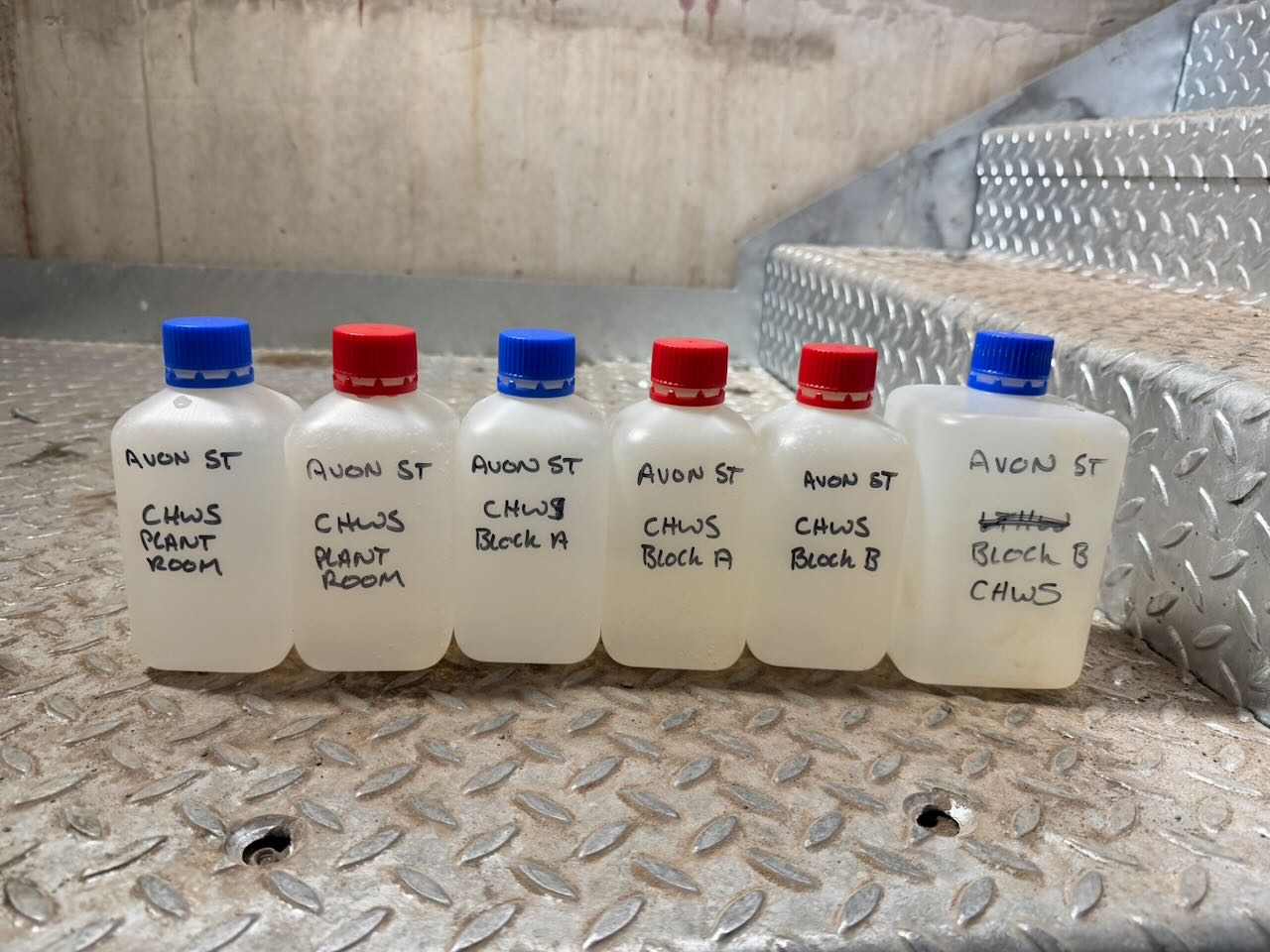 Six labeled plastic bottles with blue and red caps placed on a metal floor near metal stairs.