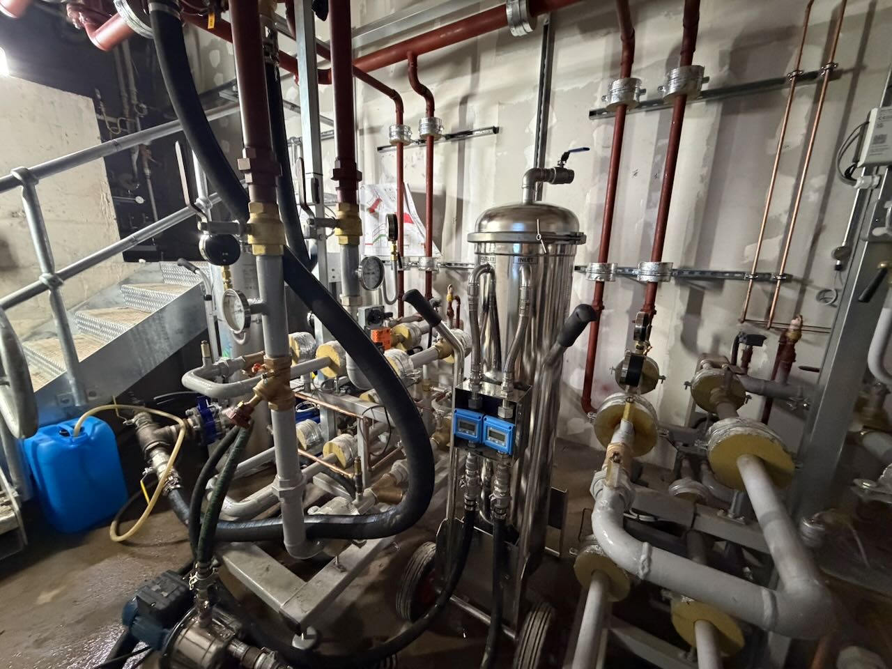 Industrial room with metal pipes, gauges, valves, a stainless steel filter, and a blue container near stairs.