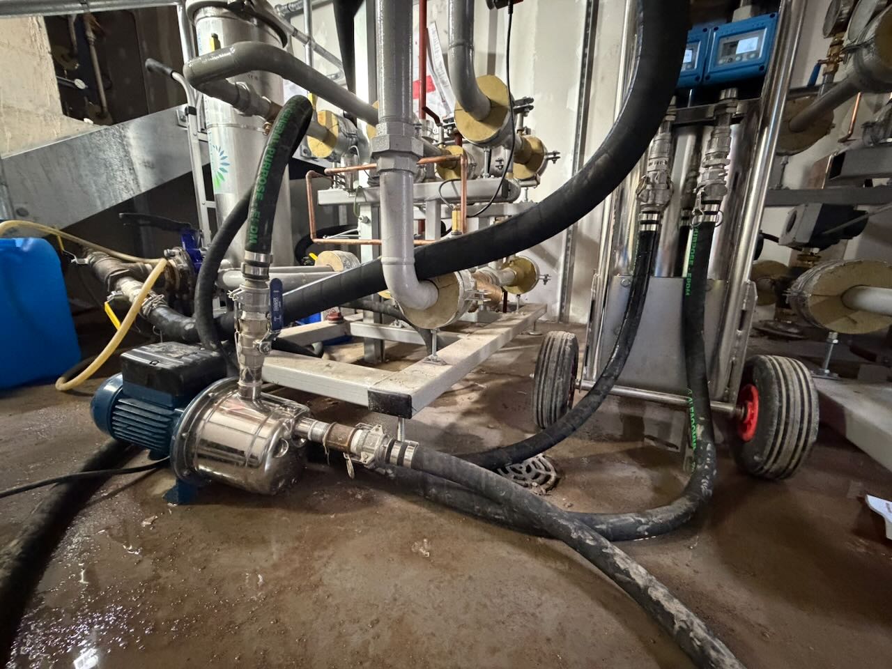 Industrial water pump with connected thick hoses and metal pipes in a wet utility room.