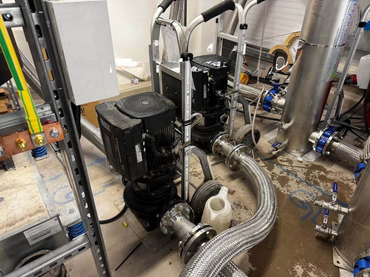 Two industrial pumps with metal flexible hoses connected in a utility room with concrete floor and valves.