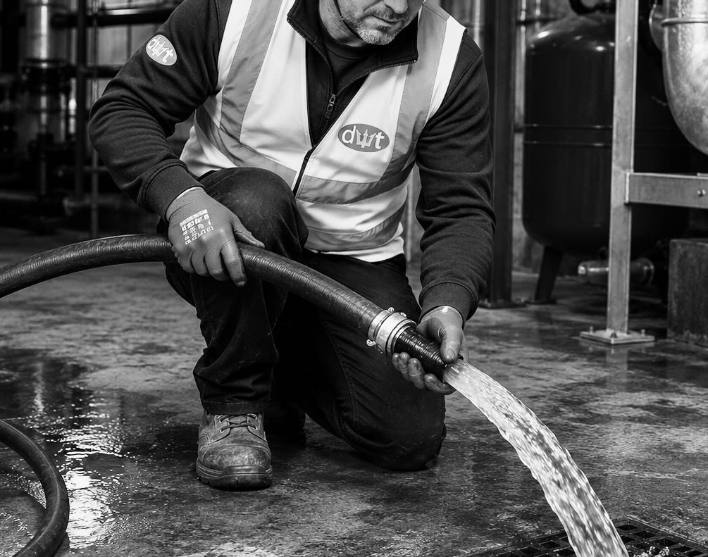 Worker wearing gloves and vest kneels while releasing water from a hose onto a floor drain.