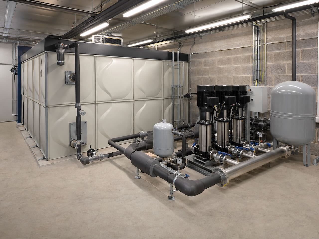 Industrial water pump system with large white tank and interconnected pipes in a concrete room.