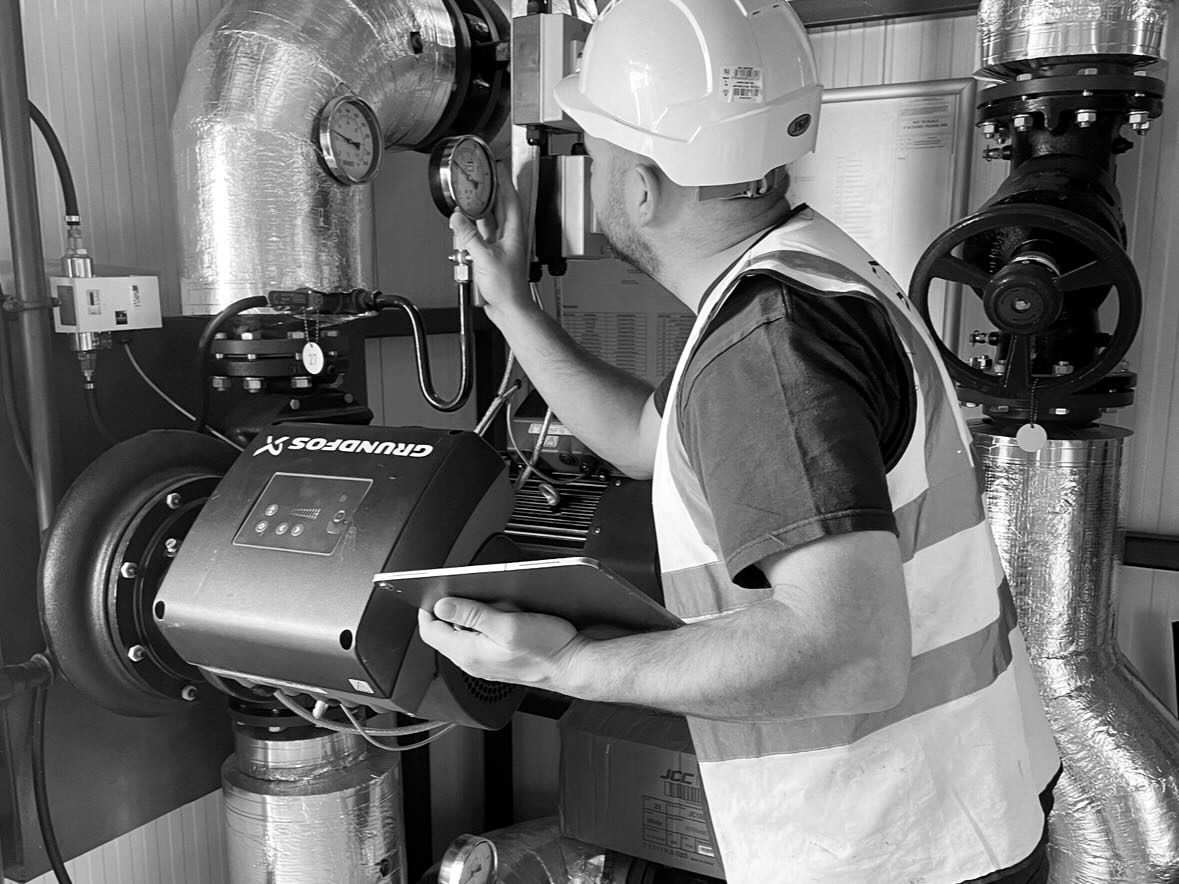 Man wearing hard hat and safety vest reading a gauge on industrial machinery and holding a tablet.