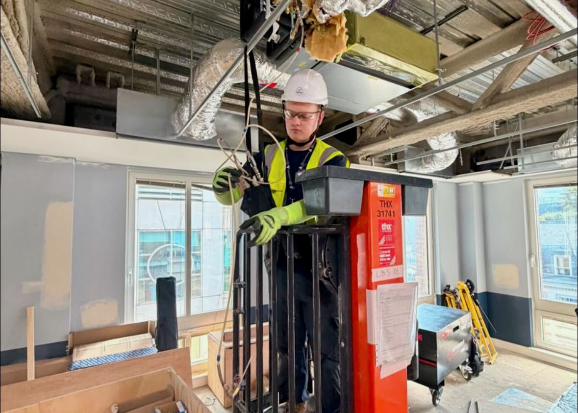 Construction worker in safety gear working on wiring while elevated on a lift inside a building.