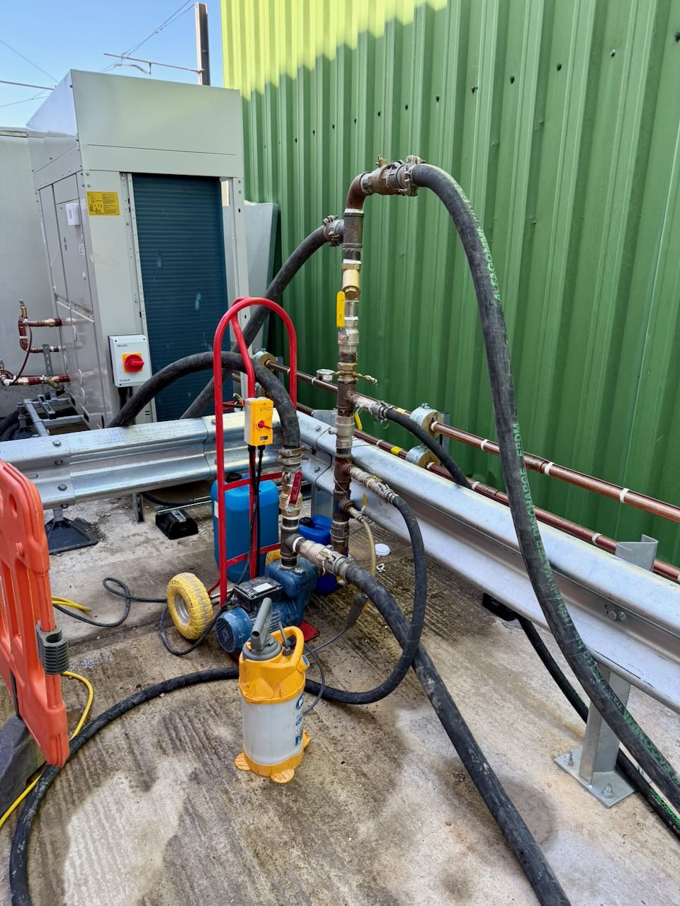 Temporary industrial pump setup with hoses, valves, and a green corrugated wall in the background.