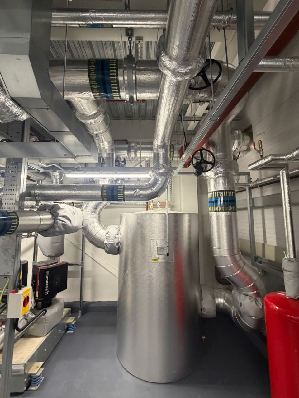 Industrial room with insulated metal pipes, valves, a large cylindrical tank, and machinery on a gray floor.