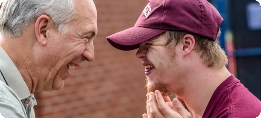 Older man and younger man with Down syndrome smiling and facing each other outdoors.