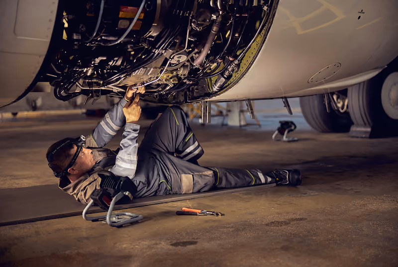 Technician lying on the ground repairing an aircraft engine inside a hangar.