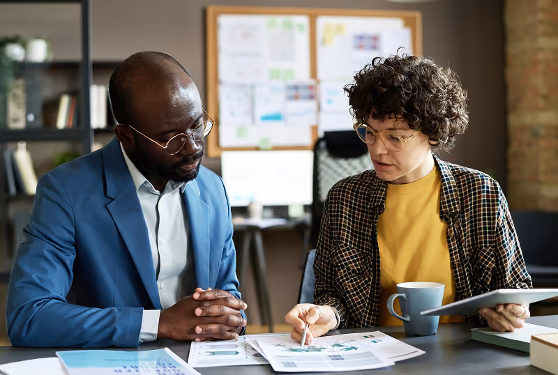 Two professionals review charts and graphs at a desk in an office setting.