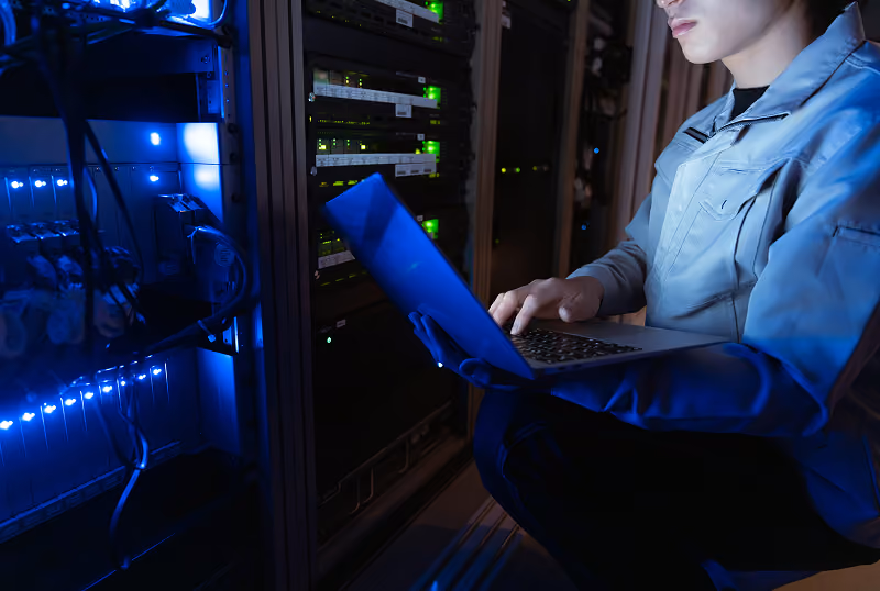 Person using a laptop inside a dimly lit server room with glowing network equipment.