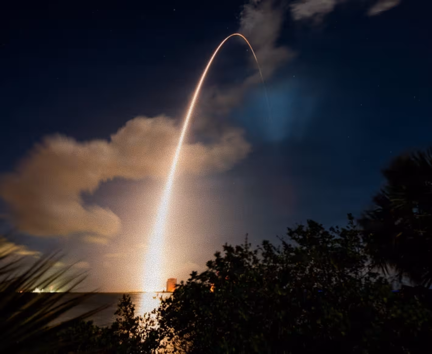 Night sky with a rocket launch creating a curved bright trail above trees and water.