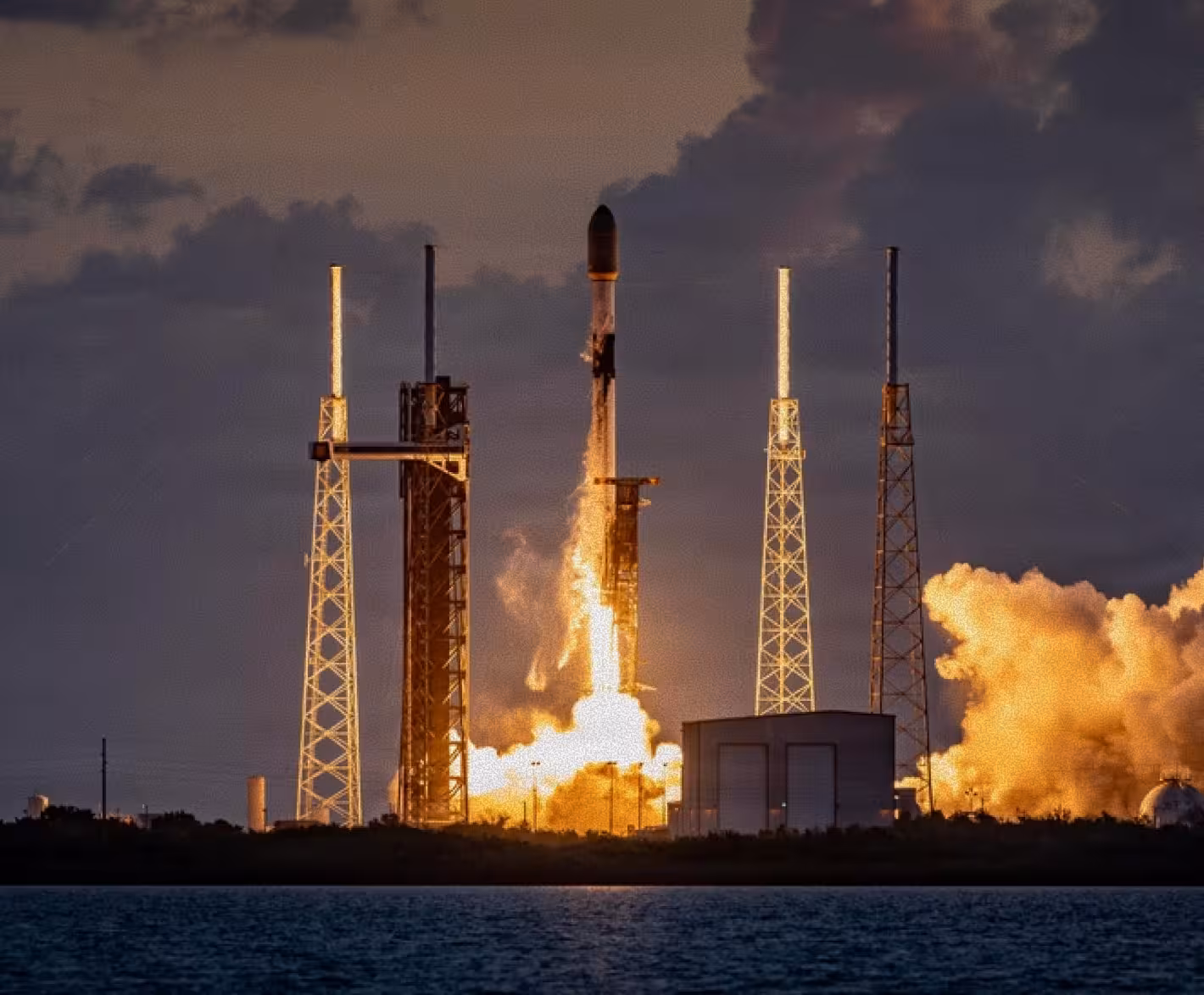 Rocket launching at dusk with flames and smoke near a body of water under cloudy sky.