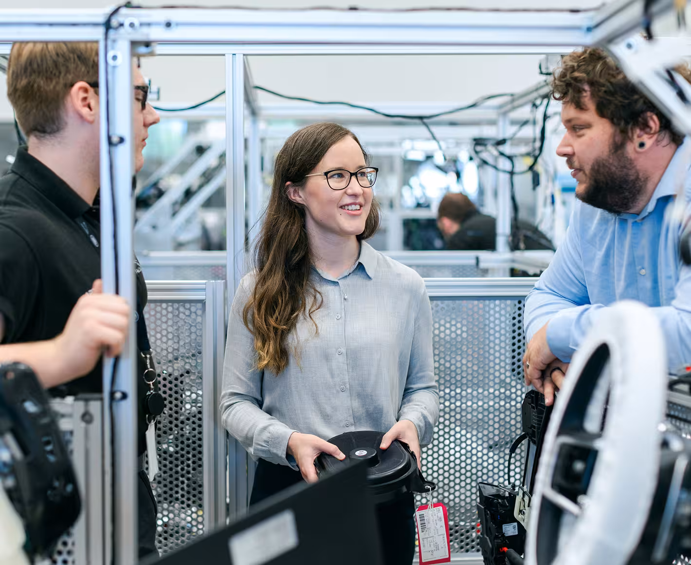 Three engineers discussing a component inside a metal-framed workstation in a lab environment.