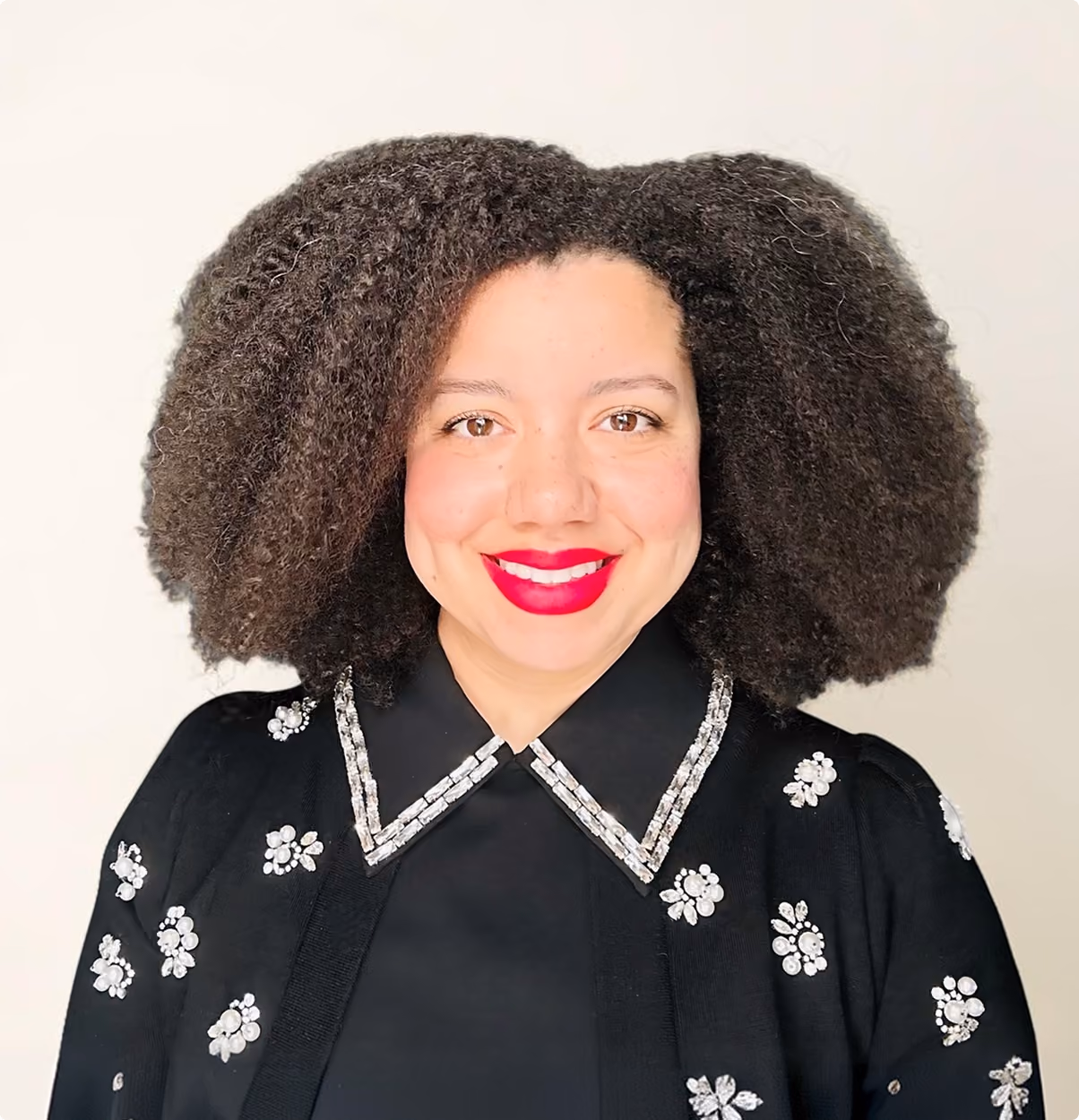 Smiling woman with voluminous curly hair wearing a black top with jeweled collar and floral designs.