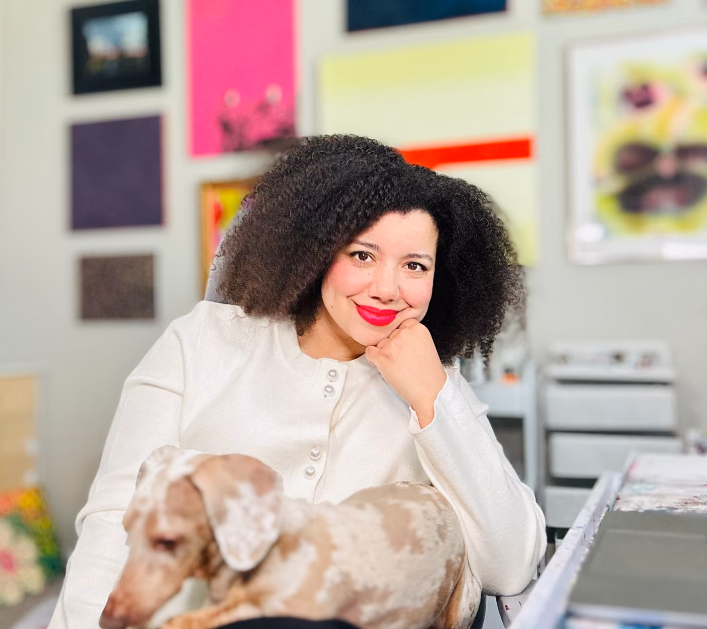 Woman with curly hair and red lipstick sitting with a small dog on her lap in a room with colorful wall art.