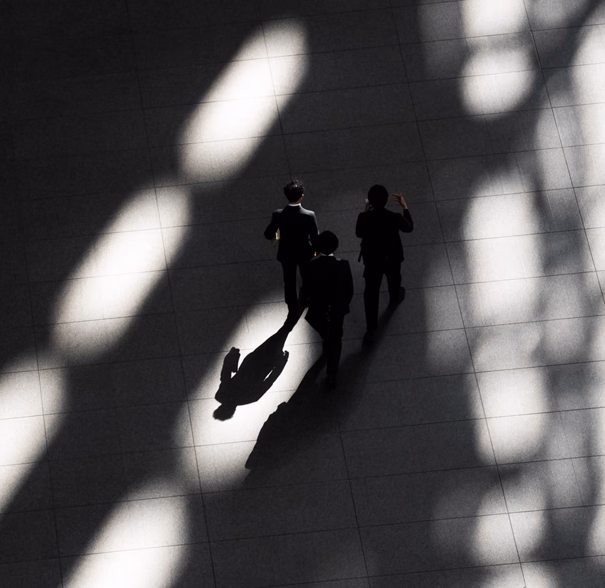 Overhead view of three people walking on a tiled floor with patterned light and shadows.
