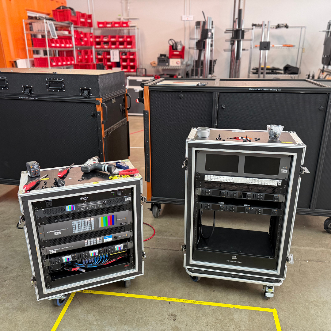 Broadcast production equipment racks and large rolling road cases staged in a production warehouse, with video switchers, monitors, cables, and tools visible.